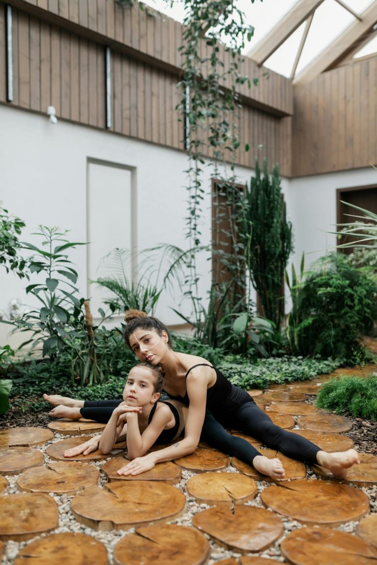 A mother and daughter doing yoga stretches indoors surrounded by plants.